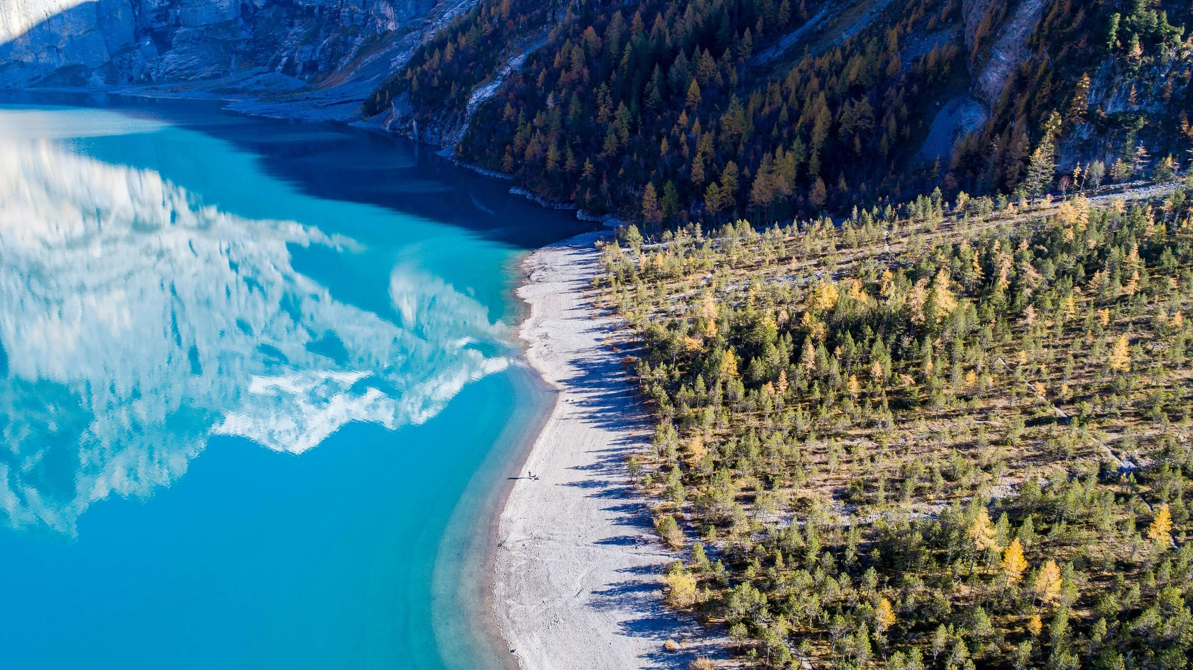 空拍視角下，一座青綠色的高山湖泊與森林相連，岸邊矗立著秋季金黃色的落葉松