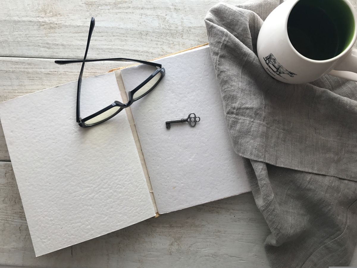 Open blank journal with reading glasses, a small key, and a cup of tea on a white wooden table